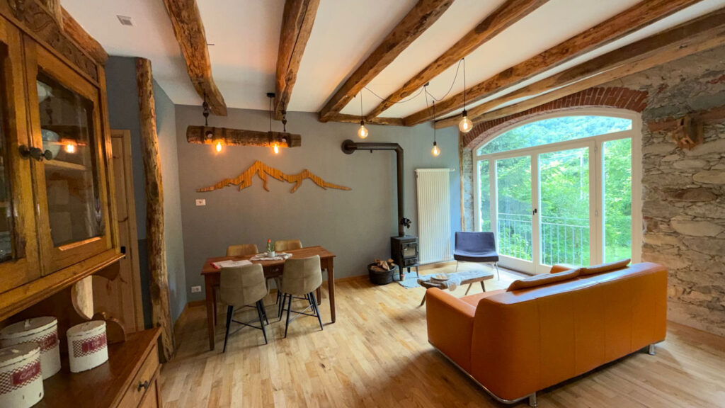 Sfeervolle woonkamer van Appartement Stella in Valsesia, Piemonte met houten balken, houtkachel en groot raam met uitzicht.